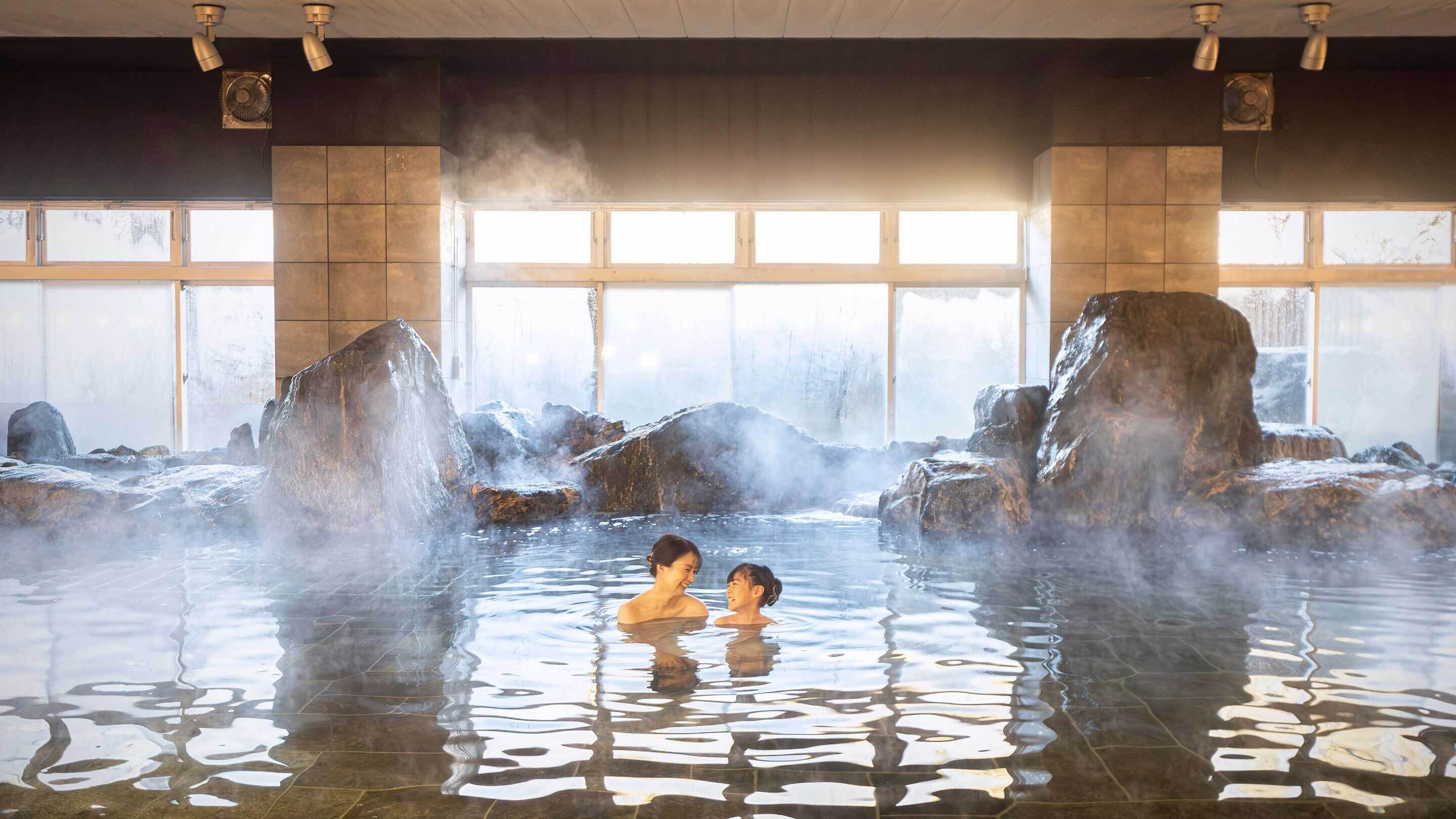 A hot spring bath with large rocks and steam, surrounded by tiled walls and large windows.