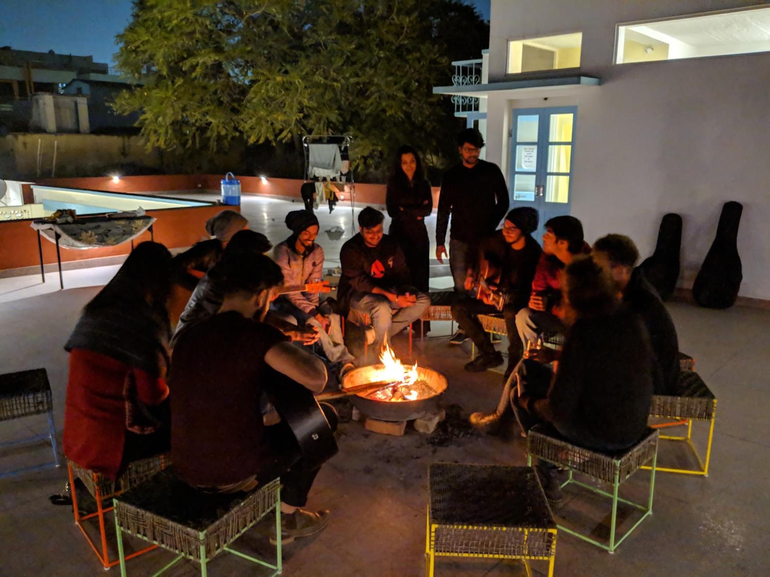 A group of people gathered around a fire pit on an outdoor patio with chairs and a table, with buildings in the background.