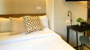 Una cama bien tendida con ropa blanca y almohadas estampadas, una mesita con una planta y un televisor montado en la pared.