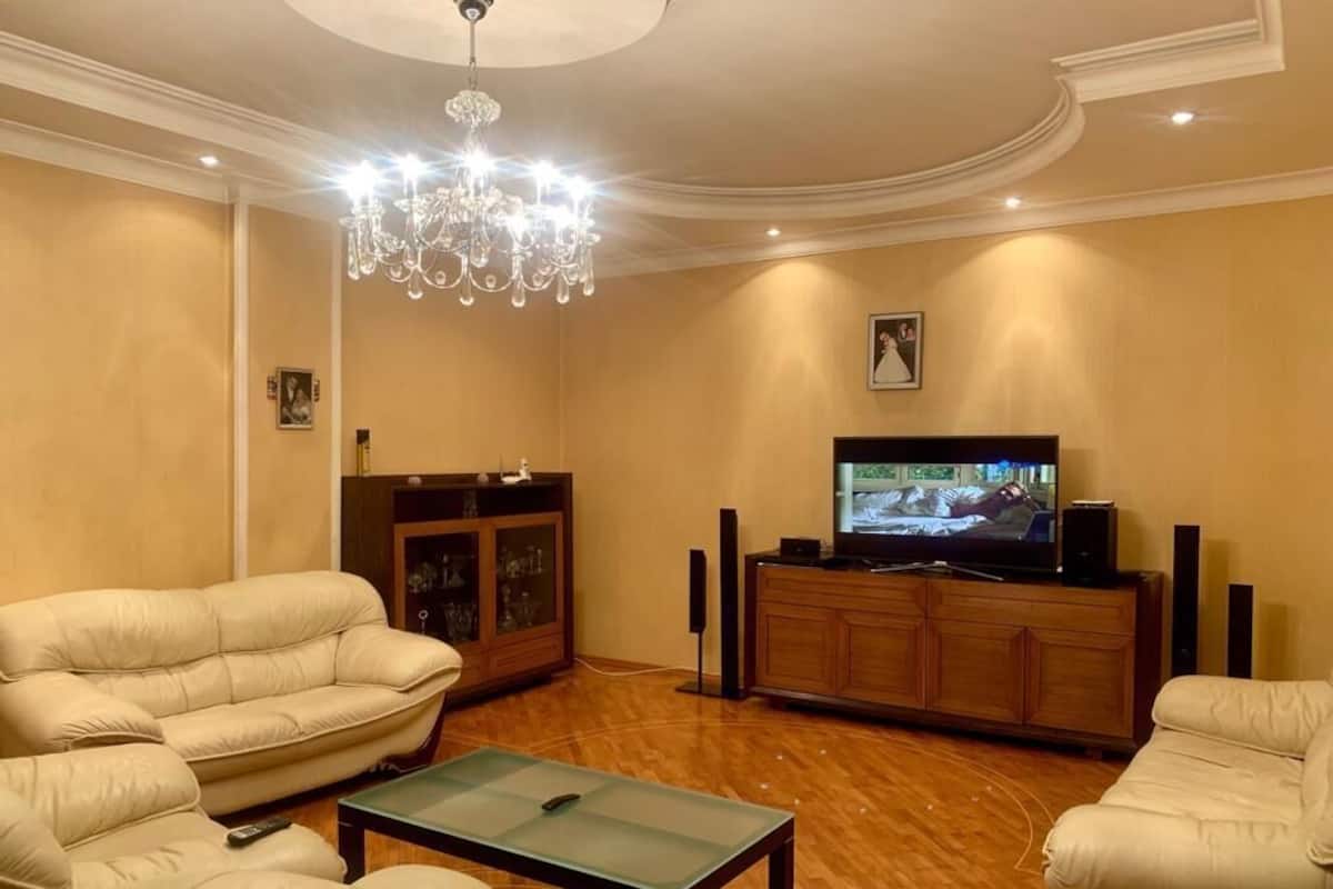 Una sala de estar con televisor, sofás de cuero, una mesa de centro de vidrio y una araña.