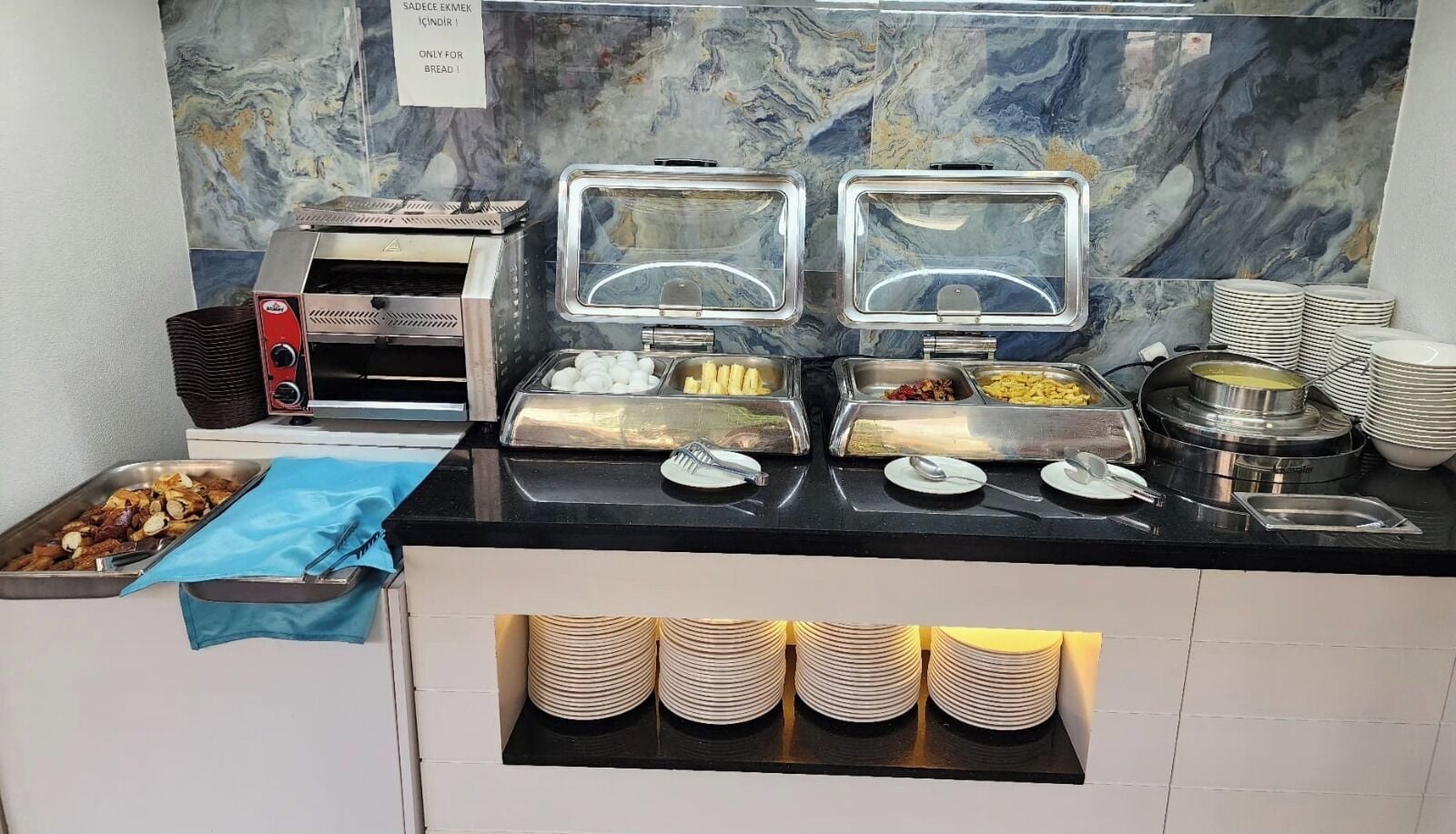 A buffet setup with various food items, a bread machine, and stacked plates.