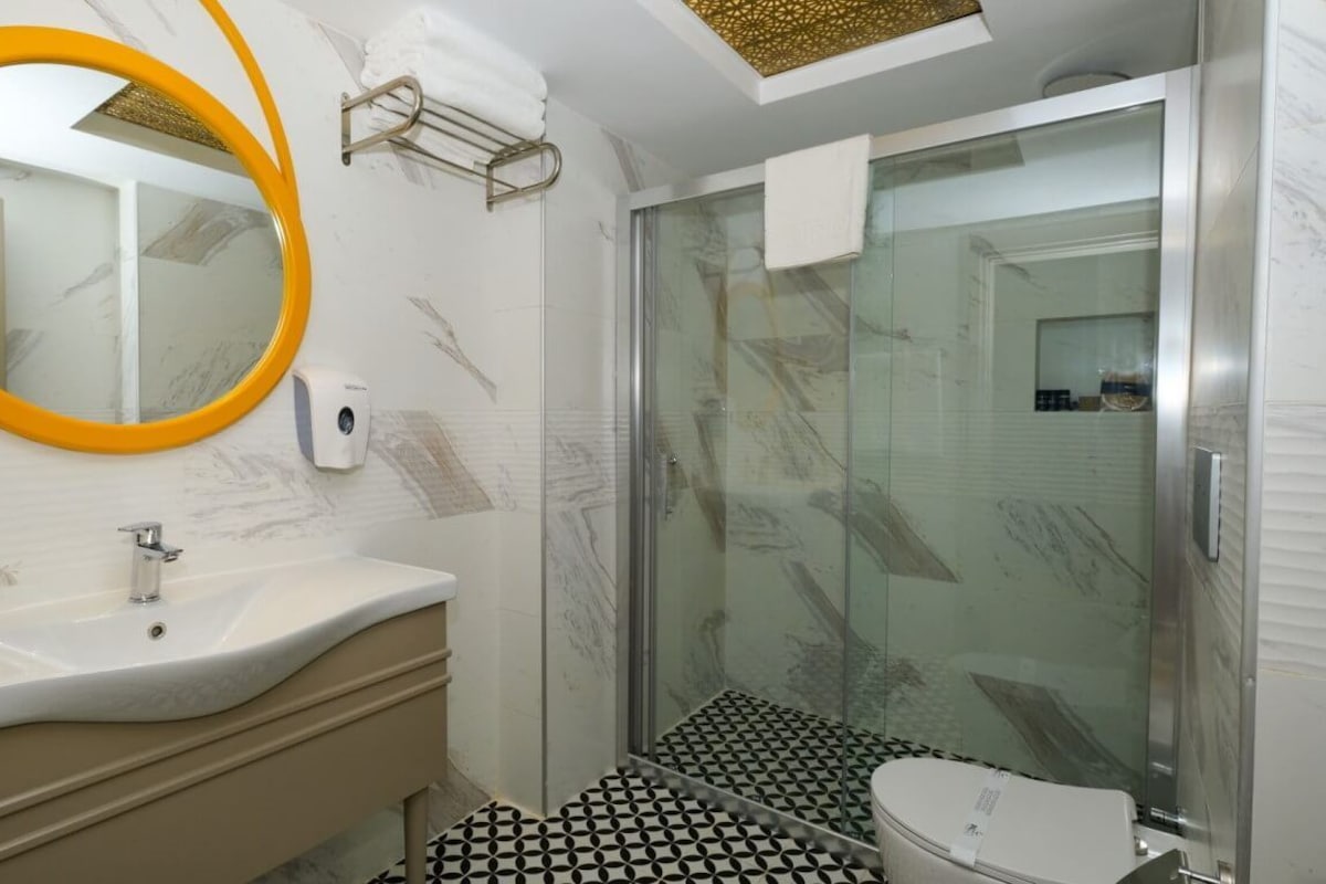 Un baño con tocador de mármol, espejo con marco amarillo, ducha con cerramiento de vidrio y piso de diseño blanco y negro.