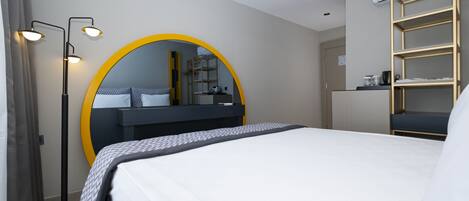 A modern hotel room with a large bed, a round mirror with a yellow frame, a bedside table, a wardrobe with a built-in shelf, and a floor lamp.