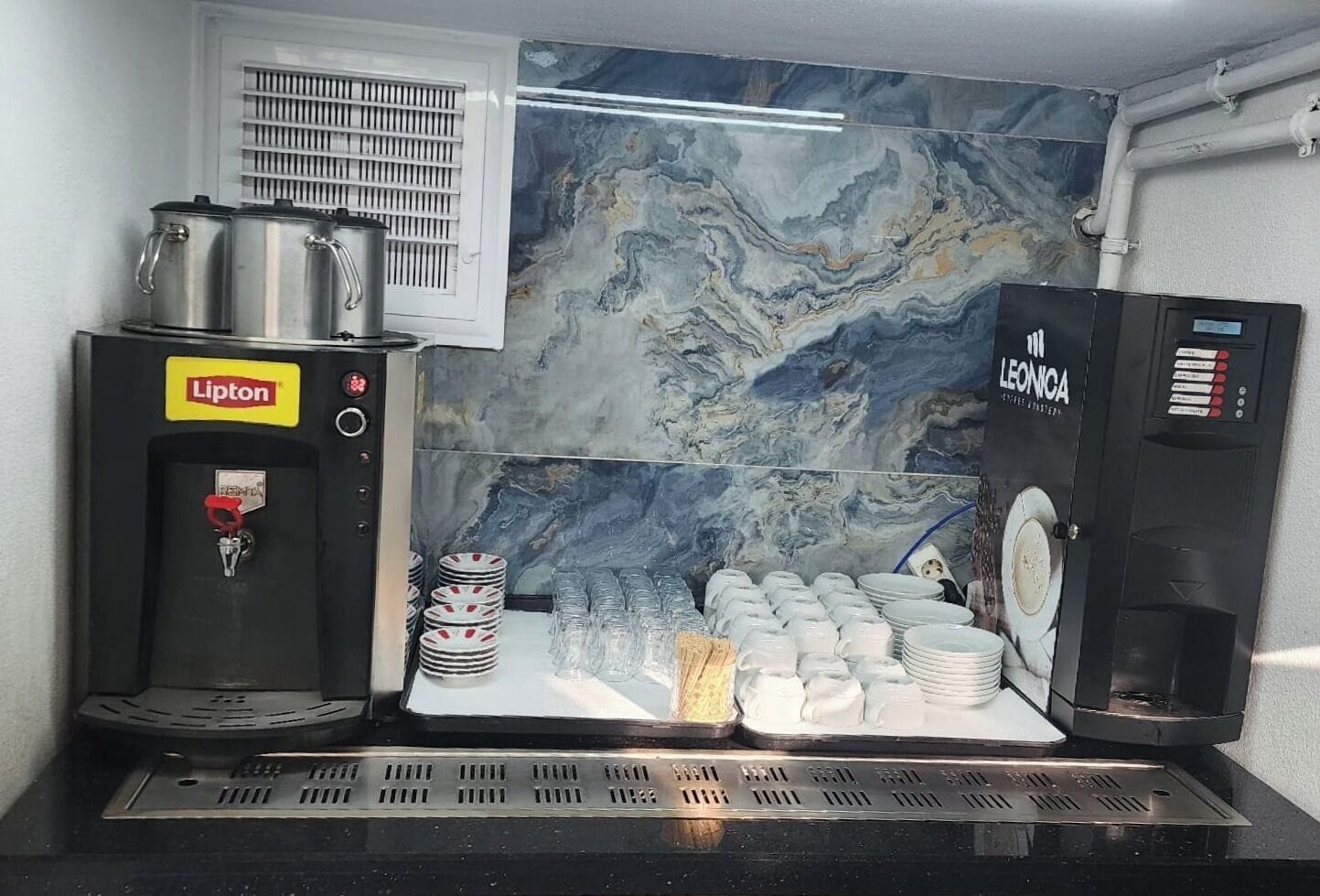 A coffee machine with a Lipton logo, a Leonica coffee machine, and a stack of white plates and glasses on a counter.
