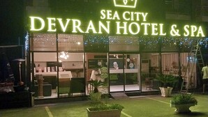 The exterior of the Sea City Devran Hotel & Spa at night, featuring a prominent crown logo, illuminated signage, and potted plants in the foreground. - Sea City Devran Hotel & Spa (Konyaalti)