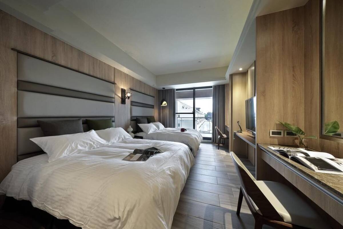 A modern hotel room with two beds, a large window, a desk with a mirror, and a TV.