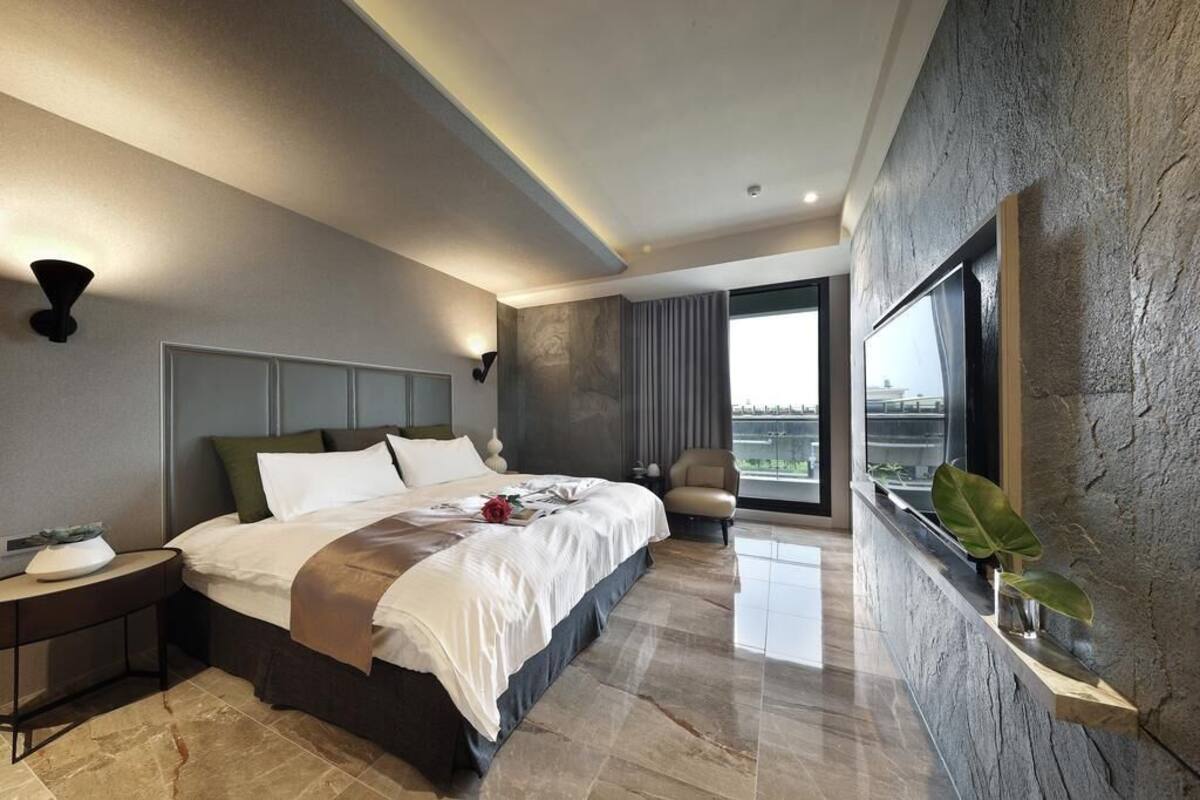 A modern hotel room with a large bed, a nightstand, a chair, a window with a view, and a plant on a shelf.