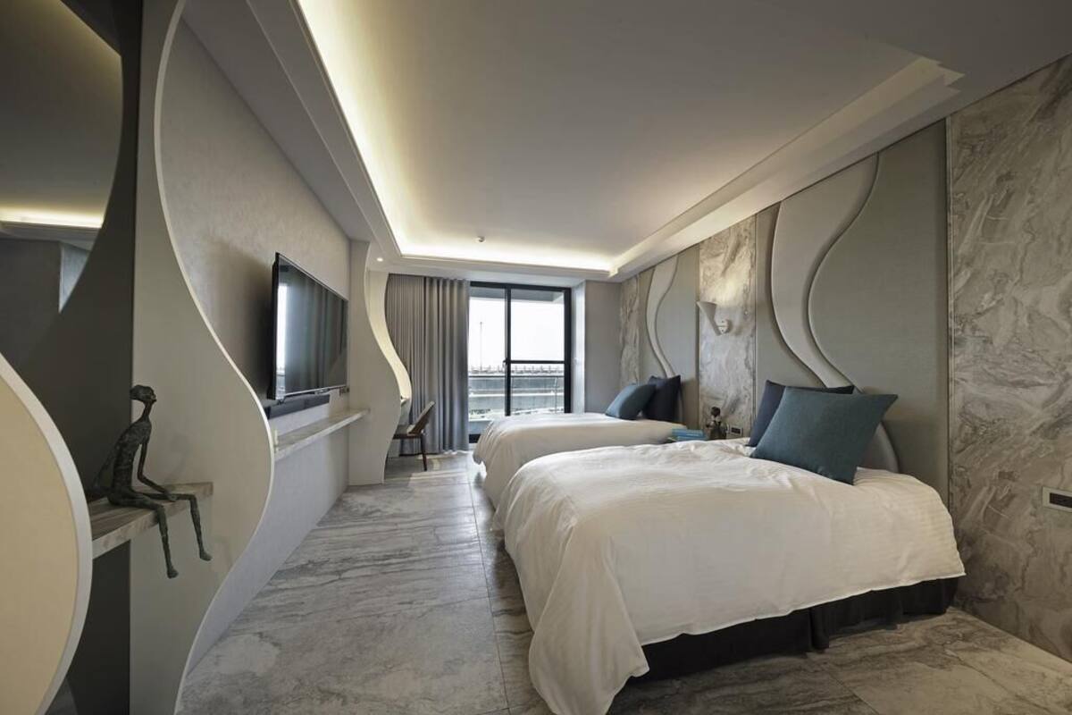 A modern hotel room with two beds, a flat-screen TV, a seating area with a chair, and a large window offering a view.