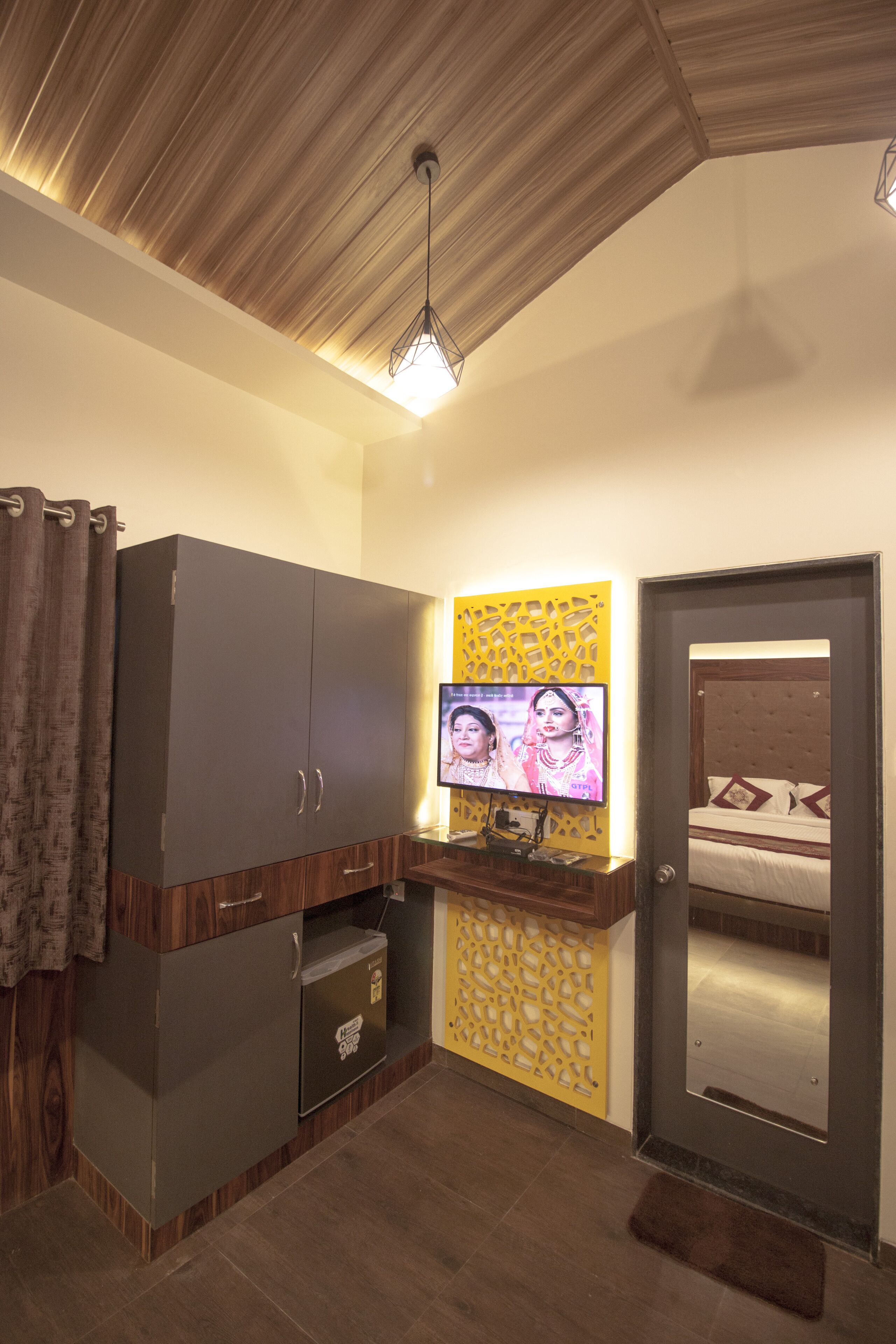 A compact room with a wardrobe, a small fridge, a TV mounted on a yellow wall, and a bed with a headboard.