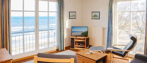 Une pièce dotée d’une grande fenêtre donnant sur la mer, d’un téléviseur, d’une table basse en bois et de deux chaises.