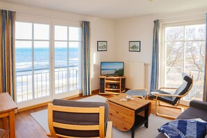 Una habitación con un ventanal grande con vista al mar, un televisor, una mesa de centro de madera y dos sillones.