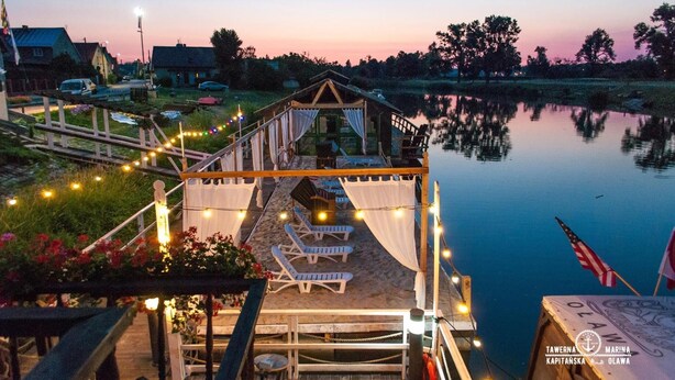 Une terrasse en bois, dotée de chaises longues et illuminée de guirlandes lumineuses, qui offre une vue sur une étendue d’eau calme.