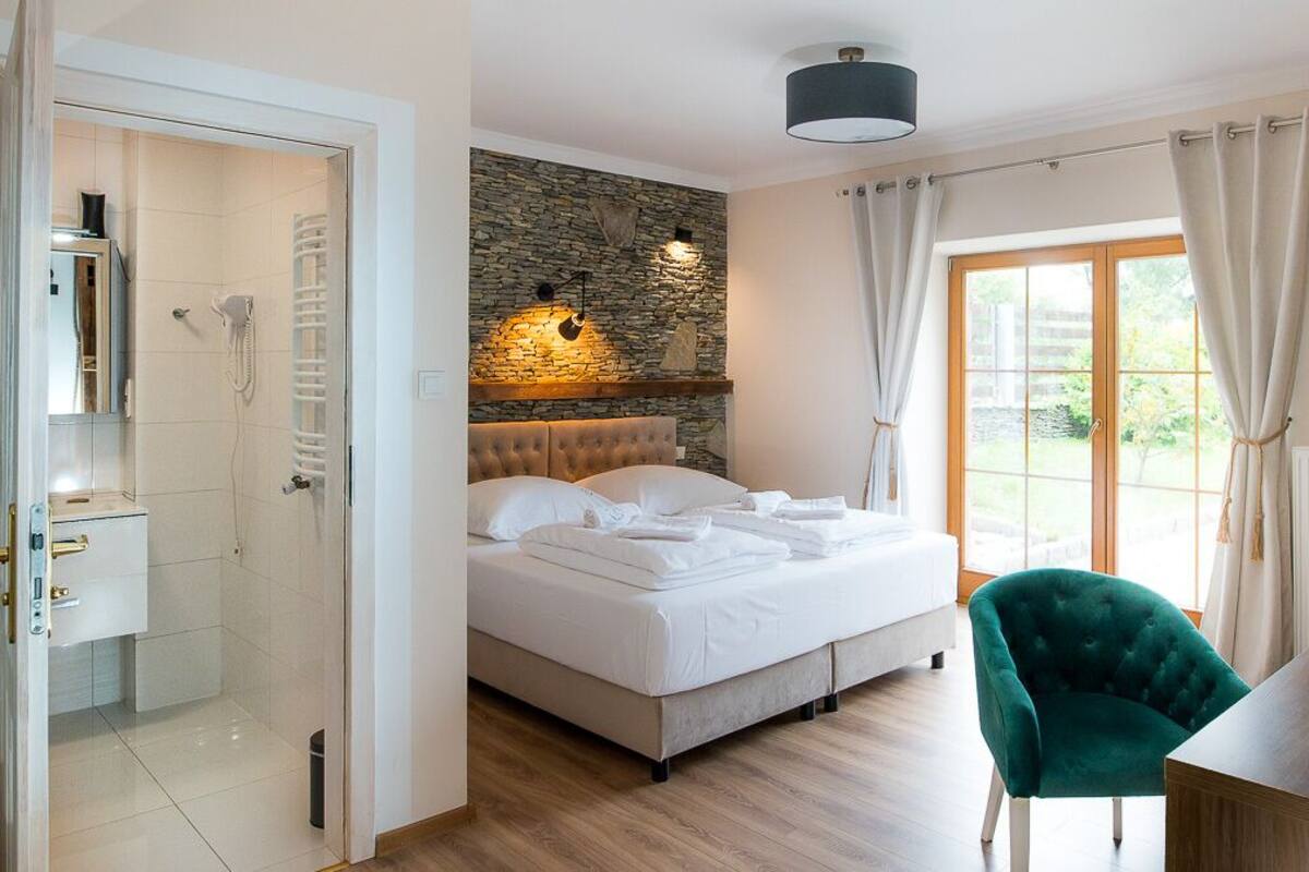 Un dormitorio con pared de piedra, una cama, una silla verde, una mesa de madera y un baño con ducha e inodoro.