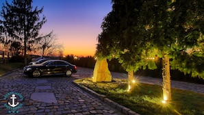 Une voiture est garée sur une allée pavée, une grande sculpture en pierre se trouve au centre, et des arbres illuminés se dressent sur la droite.