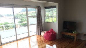 A spacious room with a wooden floor, a large sliding glass door leading to a balcony, a red bean bag chair, and a television on a wooden stand. - Come and Relax @ Rose's Retreat (with WIFI) (Whangarei Heads)