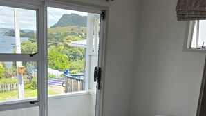 A bedroom with two beds, a balcony with a view of the sea and hills, and a window with a curtain. - Come and Relax @ Rose's Retreat (with WIFI) (Whangarei Heads)