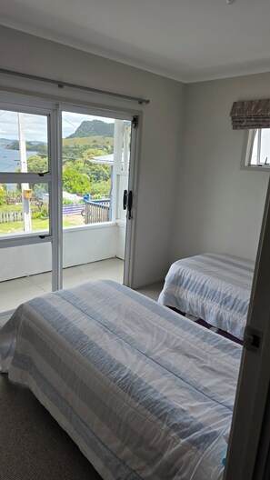 A bedroom with two beds, a balcony with a view of the sea and hills, and a window with a curtain.
