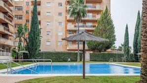 Una piscina con sombrilla de paja, rodeada de césped cuidado y palmeras, con un edificio de varios pisos al fondo.