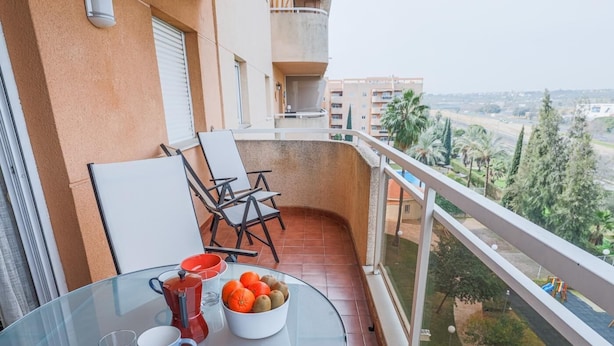 Una mesa de vidrio con un frutero, una taza roja y una taza blanca en un balcón con vistas a un parque y edificios.