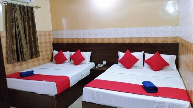 A hotel room with two beds, each with white bedding and red pillows. There is a nightstand with a phone and a lamp between the beds. The room has a patterned wall and a curtain on the left side. - Hotel Bharat Palace (Jaipur)