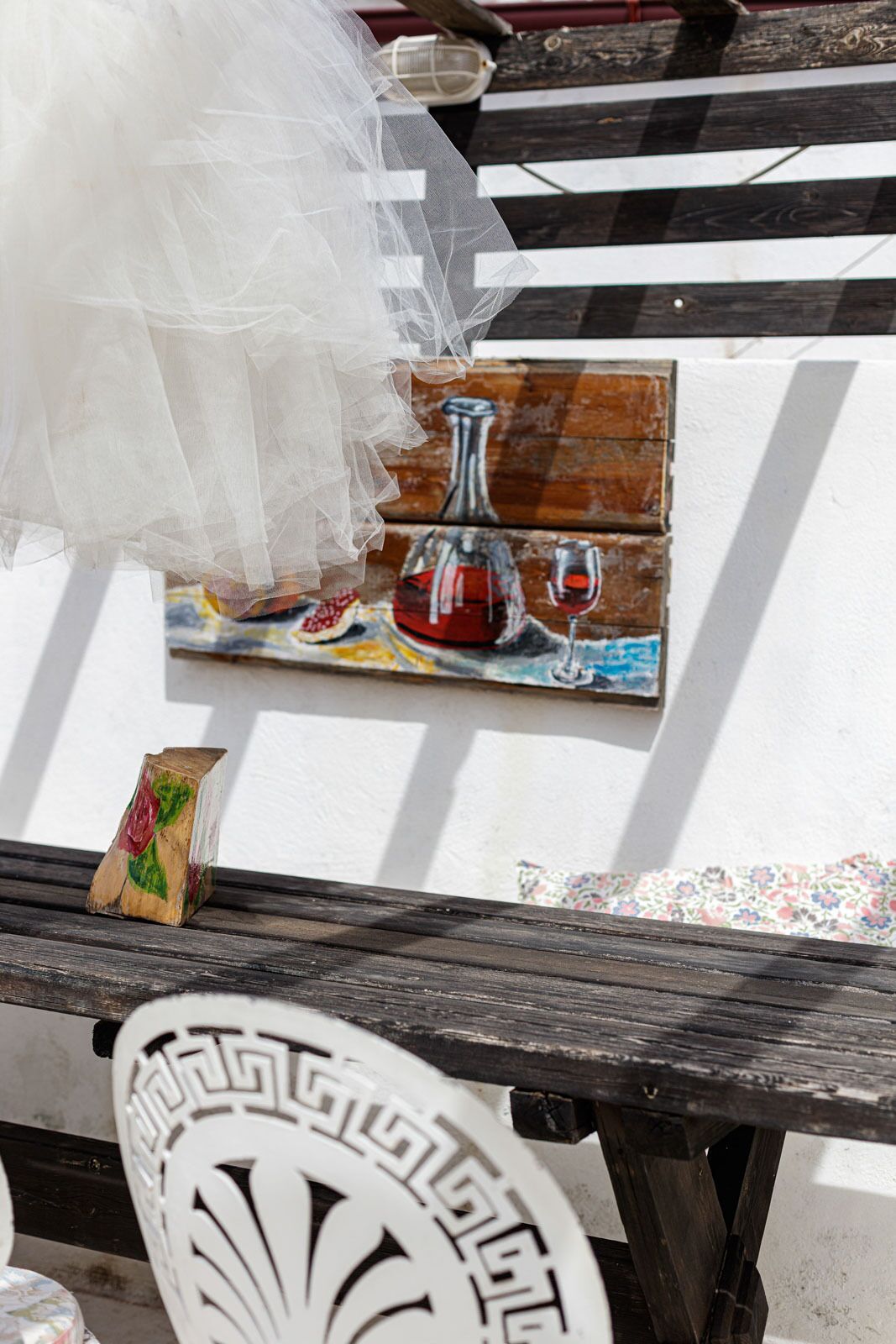 A wooden bench with a white decorative backrest, a small wooden block, and a painting of a wine bottle and glass on the wall.