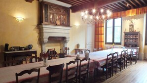 Een eetkamer met een lange tafel, houten stoelen, een kroonluchter, een open haard en een raam met gordijnen.