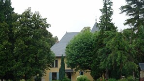 Een gebouw met twee verdiepingen, een grijze dak en een gele gevel, omringd door weelderige begroeiing en bomen.