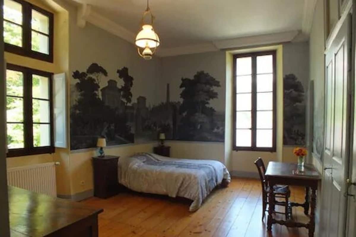 Ein Schlafzimmer mit einem Bett, einem Nachttisch, einem Tisch mit Stuhl und einem Fenster mit Blick auf Bäume.