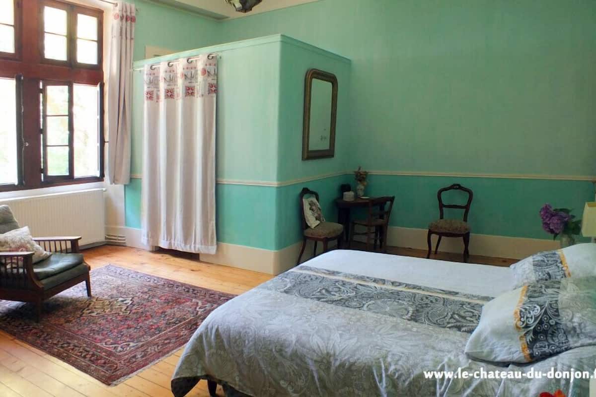 Ein Schlafzimmer mit einem Bett, zwei Stühlen, einem Tisch, einem Spiegel und einem Fenster mit Vorhängen.