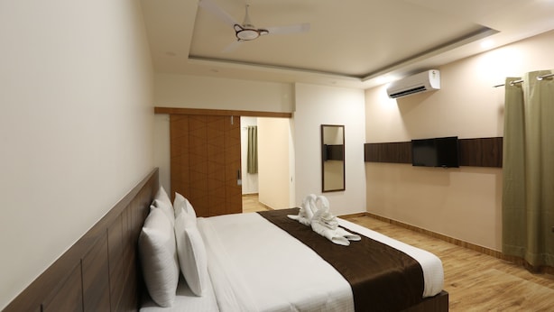 A hotel room with a bed, a ceiling fan, an air conditioning unit, a television, and a bathroom with a shower curtain. - The Ventura Grand Hotel (Bengaluru)