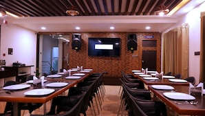 A conference room with multiple tables set for dining, a large flat-screen TV on the wall, and a sign indicating the location of the toilet. - The Ventura Grand Hotel (Bengaluru)
