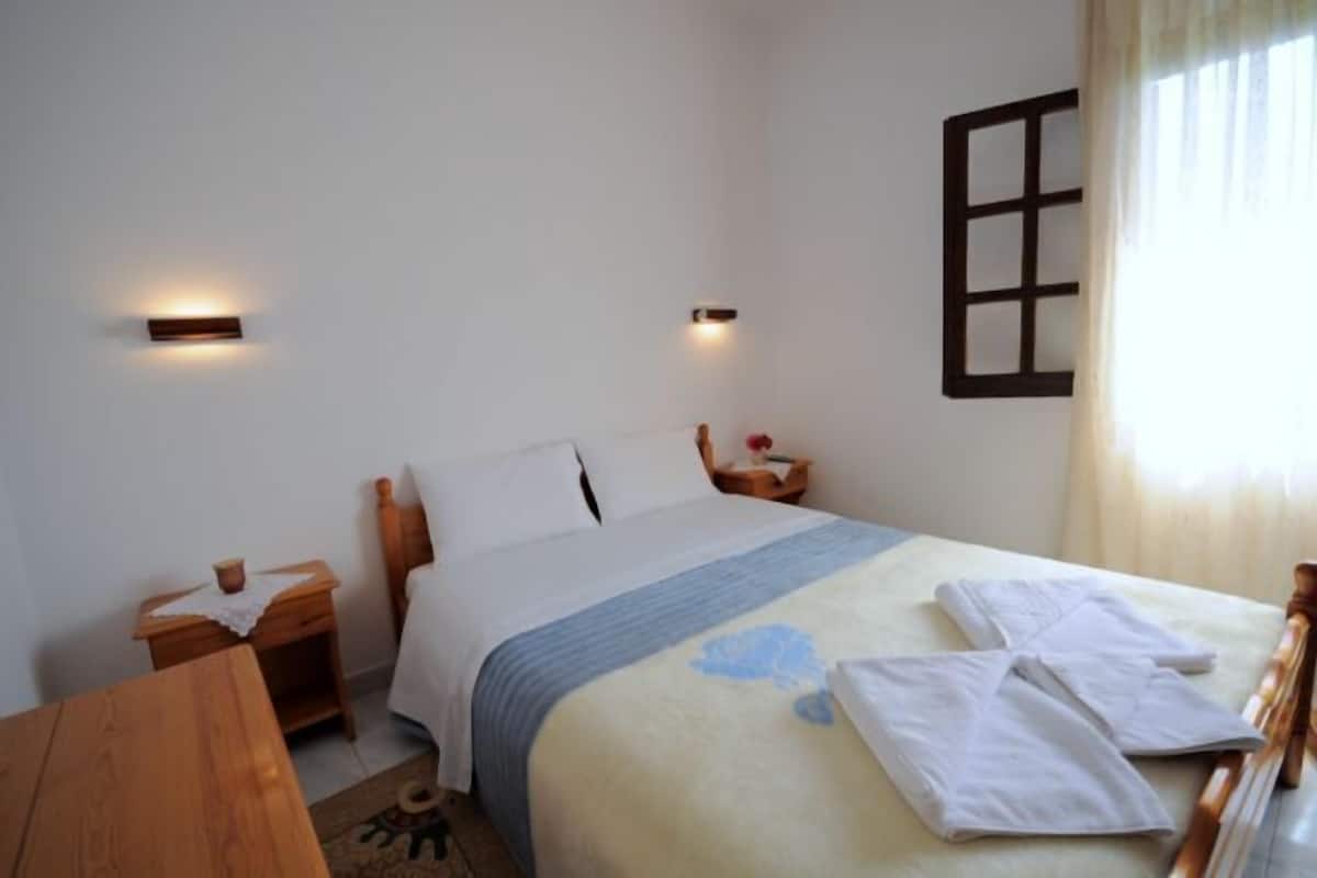 Een keurig opgemaakt bed met witte lakens en een blauw accent, een houten nachtkastje met een lamp, en een raam met houten kader.