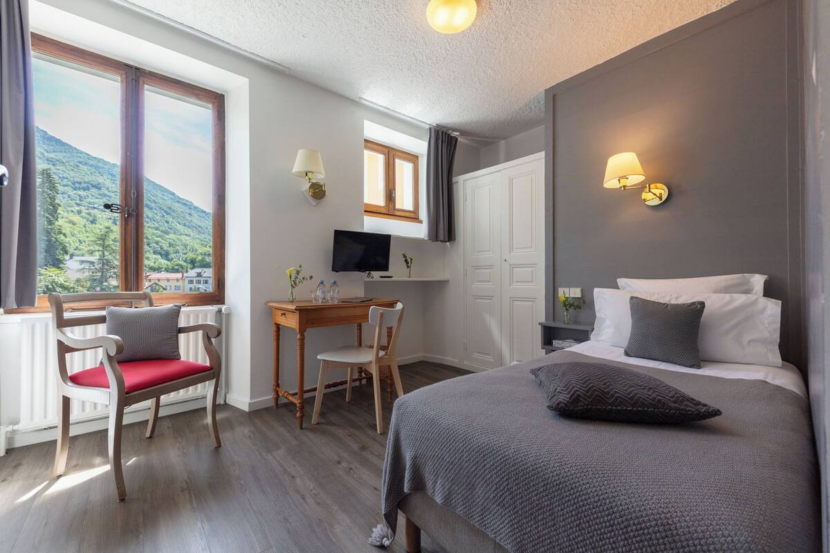 A hotel room with a bed, a desk, a chair, a TV, and a window with a view of a mountain.
