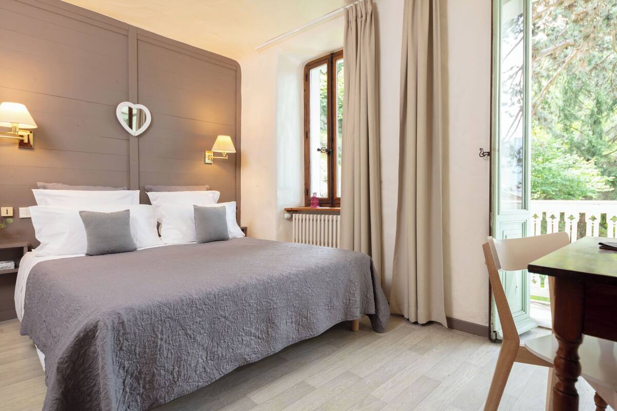 A bedroom with a large bed, two bedside tables with lamps, a window with curtains, and a view of greenery outside.