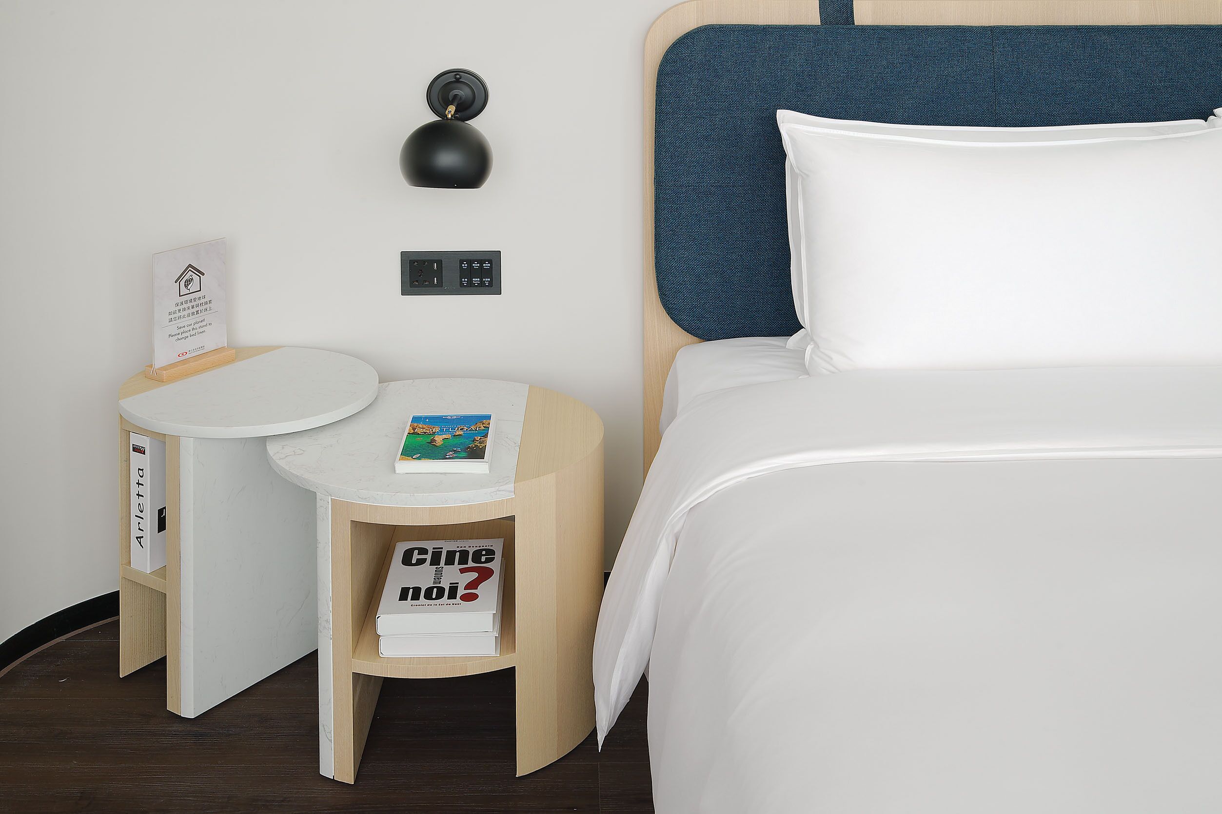 Una habitación de hotel con una cama, una mesita de noche, una lámpara y un estante pequeño con libros.
