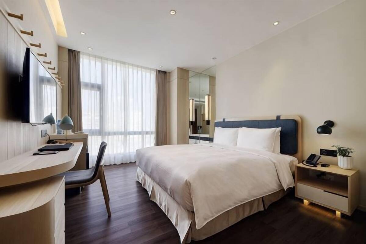 A hotel room with a bed, desk, chair, bedside tables, a TV, and a window with curtains.