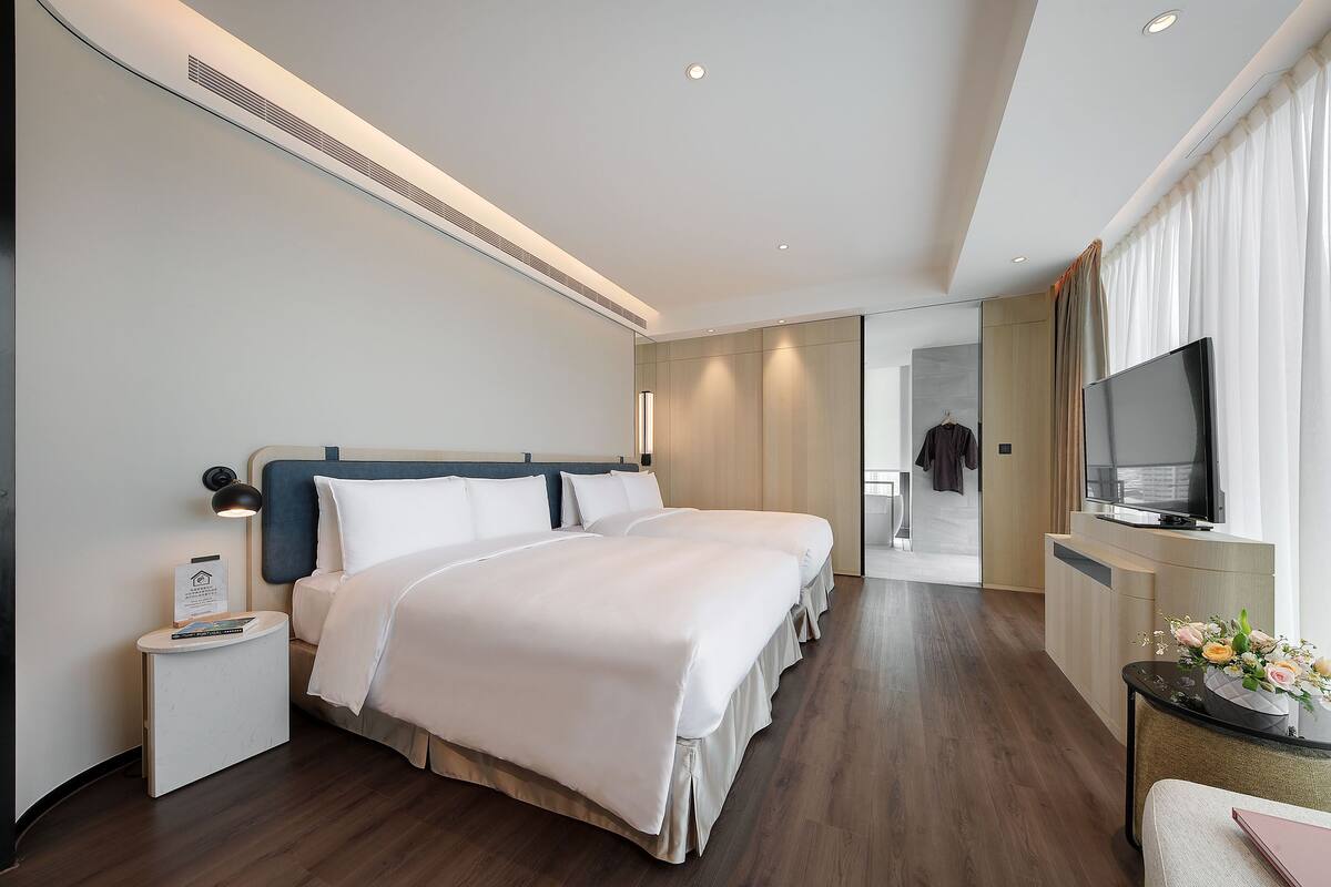 A modern hotel room with a large bed, bedside tables, a TV, and a desk with a flower arrangement.