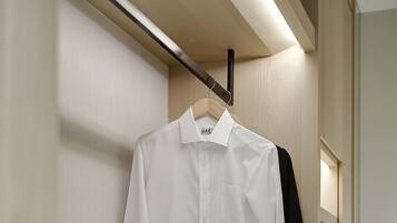 Une chemise blanche pendue à un cintre en bois, une valise grise posée sur une étagère blanche et une veste noire sur un porte-manteau en bois.