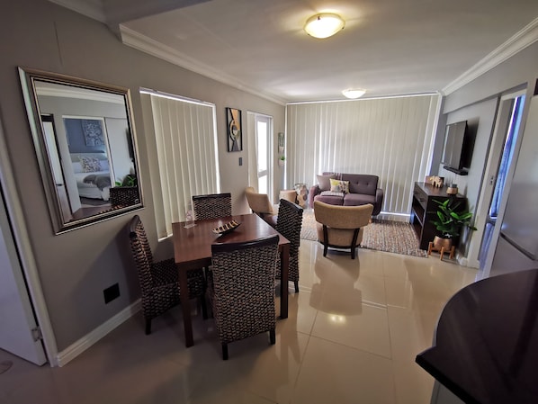A modern living room with a dining area, featuring a sofa, chairs, a coffee table, and a TV mounted on the wall.