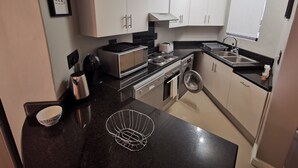 A modern kitchen with white cabinets, a black countertop, a microwave, a toaster, a kettle, and a washing machine. - The Sands B32 (Cape Town)