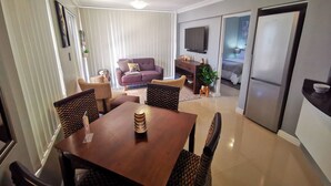A modern living room with a sofa, dining table, and chairs. There is a television mounted on the wall and a refrigerator in the kitchen area. - The Sands B32 (Cape Town)