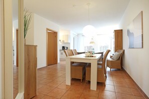 A dining area with a white table and wicker chairs, a kitchen in the background, and a large window providing natural light.