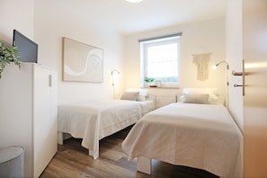 A room with two beds, a large window, a plant, and a painting.