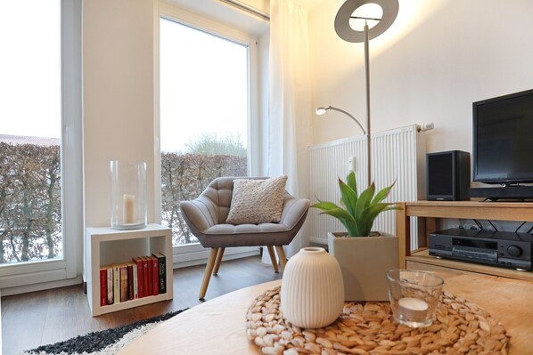 A modern living room with a sofa, a side table with books, a floor lamp, a television, and a view of the outdoors.