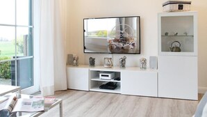 A modern living room with a large flat-screen TV mounted on a white cabinet, a glass coffee table with magazines, and a view of a garden through a large window.