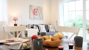 A modern living room with a sofa, a dining table with breakfast items, and a large window with a view of greenery.