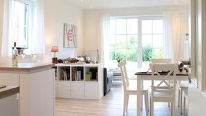 A bright, spacious living area with a dining table, chairs, and a sofa. There is a kitchen area with cabinets and a window.