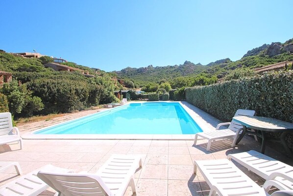 A rectangular swimming pool with white lounge chairs and a table on a tiled patio, surrounded by greenery and hills.