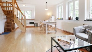 Een ruime woonkamer met een houten trap, een eettafel met stoelen, een glazen salontafel en een grijze bank.