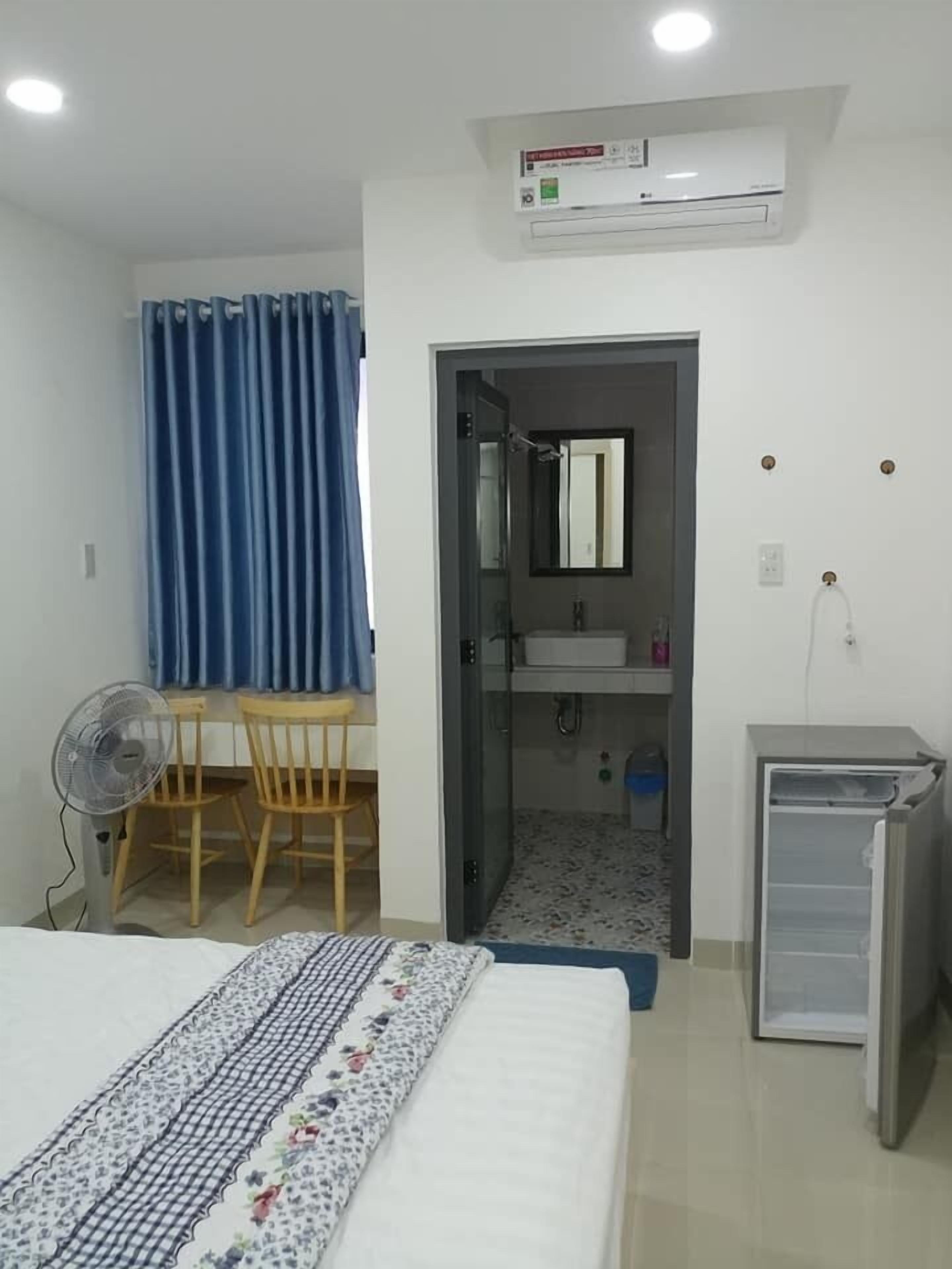 Et soveværelse med seng, skrivebord med stol, ventilator, lille køleskab og badeværelse med spejl og vask.